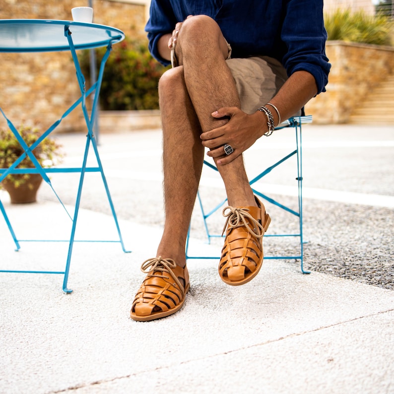 May include: A pair of light brown leather sandals with a woven design. The sandals have tan laces and are worn by a person sitting on a blue chair. The sandals are a stylish footwear choice for warm weather.