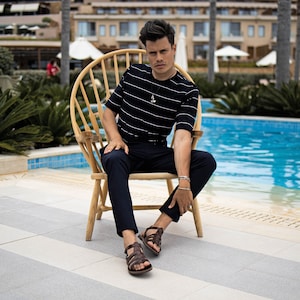 May include: A man wearing a black and white striped short-sleeve shirt, dark blue pants, and brown leather sandals sits in a wooden chair. The chair is made of light-colored wood. A silver necklace and bracelet are visible. A swimming pool is in the background.