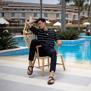 May include: A person wearing a striped navy blue and white shirt sits in a wooden chair by a pool. The person is wearing brown sandals and has their legs crossed.