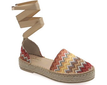 May include: Espadrille sandals with a colorful zigzag pattern in red, yellow, and brown. The sandals have a woven jute sole and beige wrap-around ankle straps. The open-sided design features a low heel and a comfortable fit.