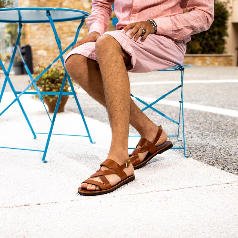 May include: A pair of brown leather sandals with buckle closures. The sandals are worn by a person sitting on a blue folding chair.