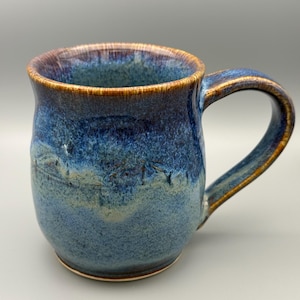 May include: A handmade ceramic mug with a blue and brown glaze. The mug has a rounded body, a curved handle, and a brown rim. The blue glaze has variations in color, creating a textured appearance. The mug is designed for hot beverages.