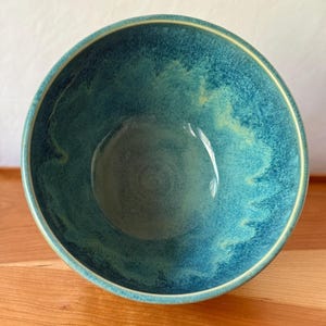 Könnte beinhalten: Eine Keramikschale mit türkisfarbener und blaugrüner Glasur. Die Schale hat eine abgerundete Form mit einem leicht ausgestellten Rand und einem gelb-goldenen Akzent. Das Innere der Schale hat ein gesprenkeltes Aussehen mit Farb- und Texturvariationen.