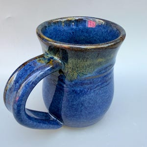 Könnte beinhalten: Eine handgefertigte Keramiktasse mit einer glänzenden, tiefblauen Glasur. Die Tasse hat einen gebogenen Henkel und einen kontrastierenden Rand mit Braun- und Grüntönen. Der Korpus der Tasse hat ein strukturiertes Aussehen.