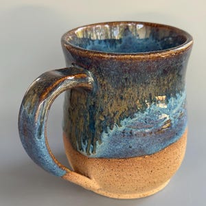 Puede incluir: Taza de cerámica hecha a mano con un esmalte único. La taza presenta un borde marrón, que transiciona a tonos azules y verdes, y una base beige. El asa también es azul y marrón. Ideal para café o té.