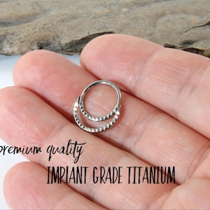 May include: A silver-coloured, double-hoop nose ring with a textured, geometric design. The text "premium quality" and "IMPLANT GRADE TITANIUM" are visible. The jewellery is held in a hand.
