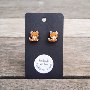 May include: Fox-shaped stud earrings on a black display card. The earrings are orange and white foxes with detailed faces. The card has a white sticker with the text "handmade with love".