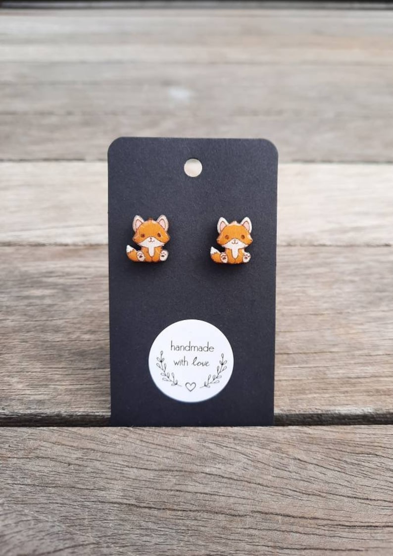 May include: Fox-shaped stud earrings on a black display card. The earrings are orange and white foxes with detailed faces. The card has a white sticker with the text "handmade with love".