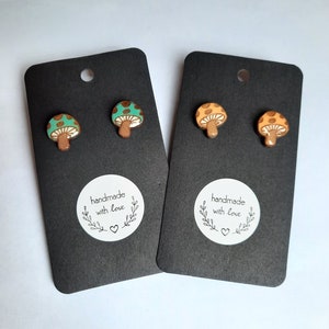 May include: Two pairs of mushroom stud earrings. One pair is green with brown spots and the other pair is orange with brown spots. The earrings are on black cards with a white circle that says "handmade with love" and a small heart.