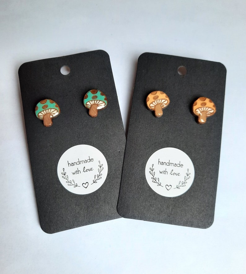 May include: Two pairs of mushroom stud earrings. One pair is green with brown spots and the other pair is orange with brown spots. The earrings are on black cards with a white circle that says "handmade with love" and a small heart.