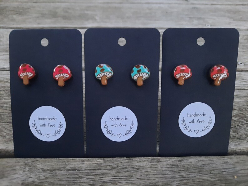 May include: Three pairs of mushroom-shaped earrings on black card stock. The earrings are red with white polka dots, turquoise with white polka dots, and red with white polka dots. Each card has a white circle with the text "handmade with love" and a small heart.