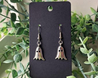 May include: A pair of silver-toned ghost-shaped dangle earrings featuring two circular cutouts for eyes. The earrings are attached to a rectangular black display card with a hole at the top for hanging, showcasing a metallic finish and smooth edges.