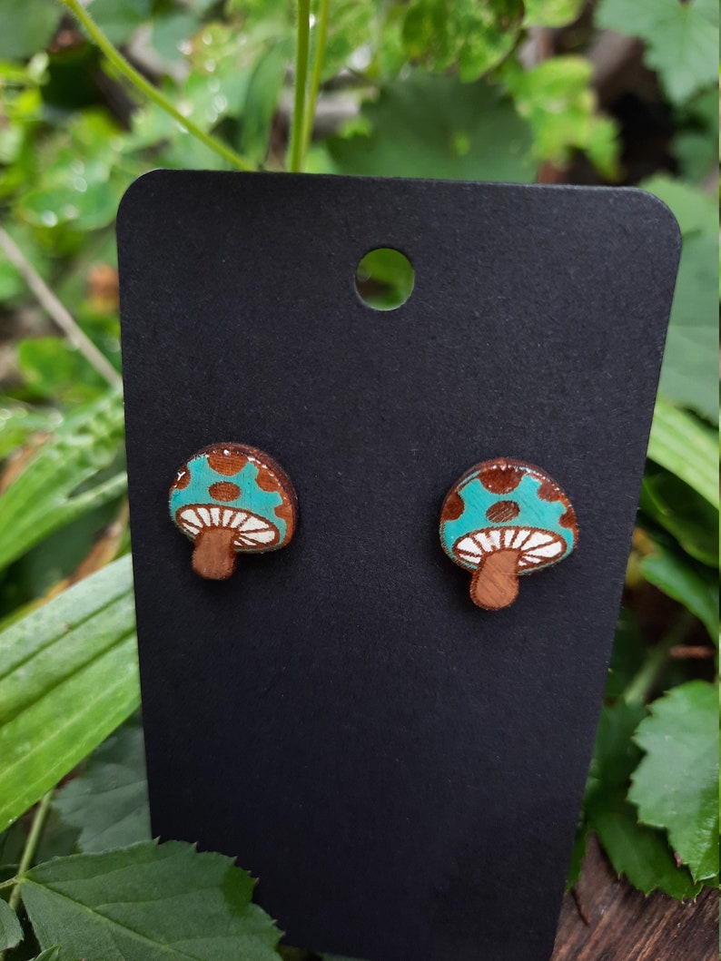 May include: A pair of wooden mushroom earrings with a teal blue and brown design. The mushrooms have white spots and are on a black card.