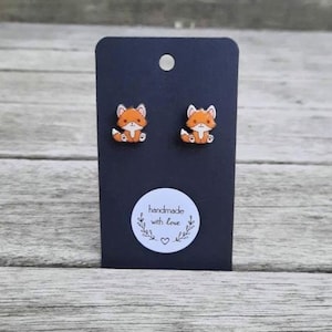 May include: A pair of orange and white fox stud earrings on a black display card. The card has a small white sticker with the text "handmade with love" and a heart. The earrings are set against a wooden background.