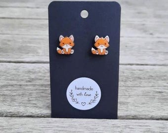 May include: A pair of orange and white fox stud earrings on a black display card. The card has a small white sticker with the text "handmade with love" and a heart. The earrings are set against a wooden background.