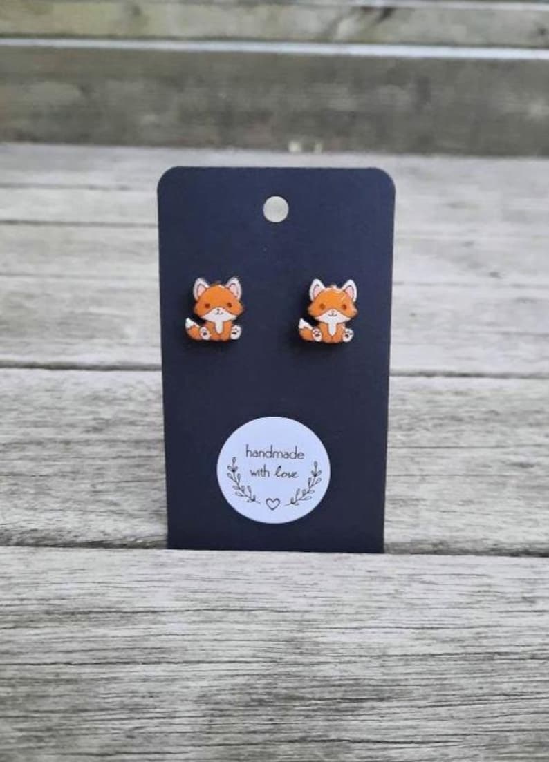 May include: A pair of orange and white fox stud earrings on a black display card. The card has a small white sticker with the text "handmade with love" and a heart. The earrings are set against a wooden background.