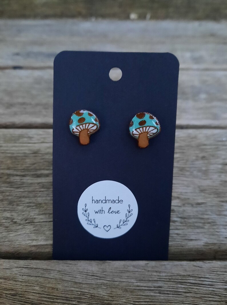 May include: A pair of mushroom-shaped earrings with brown stems and teal caps with white polka dots. The earrings are displayed on a black card with a white circle sticker that says "handmade with love".