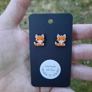 May include: Fox-shaped stud earrings displayed on a black card. The earrings depict orange and white foxes with black detailing. The card has a white sticker with the text "handmade with love" and a heart symbol.