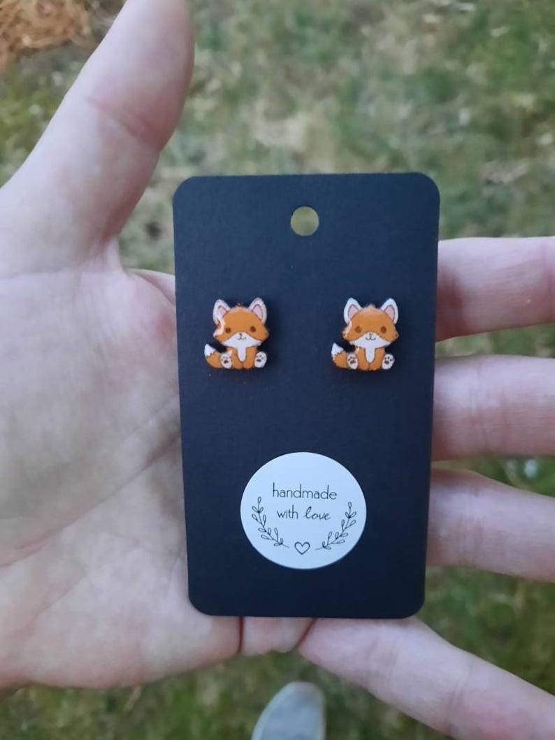 May include: Fox-shaped stud earrings displayed on a black card. The earrings depict orange and white foxes with black detailing. The card has a white sticker with the text "handmade with love" and a heart symbol.