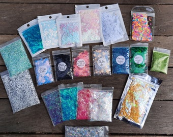 May include: Assortment of small bags filled with various colors and types of glitter. The bags are labeled with names like "Ice Queen", "Mermaid Scales", and "Flower Power".