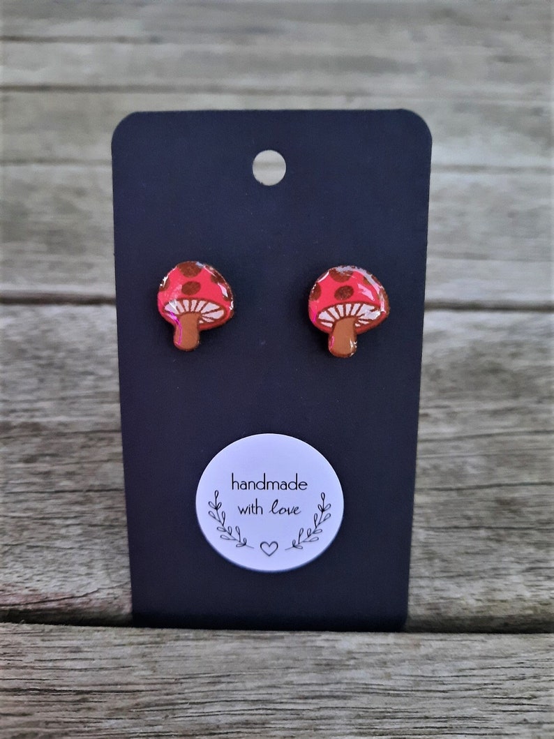 May include: A pair of red and white mushroom stud earrings on a black card with a white circle sticker that says "handmade with love" and a small heart.