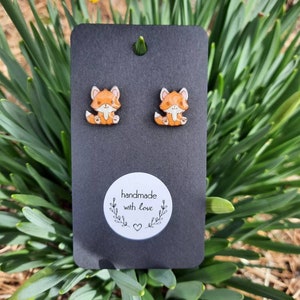 May include: Fox-shaped stud earrings on a black card. The earrings are orange and white with a cartoon design. The card has a small hole at the top and a white sticker that says "handmade with love". The background is green.