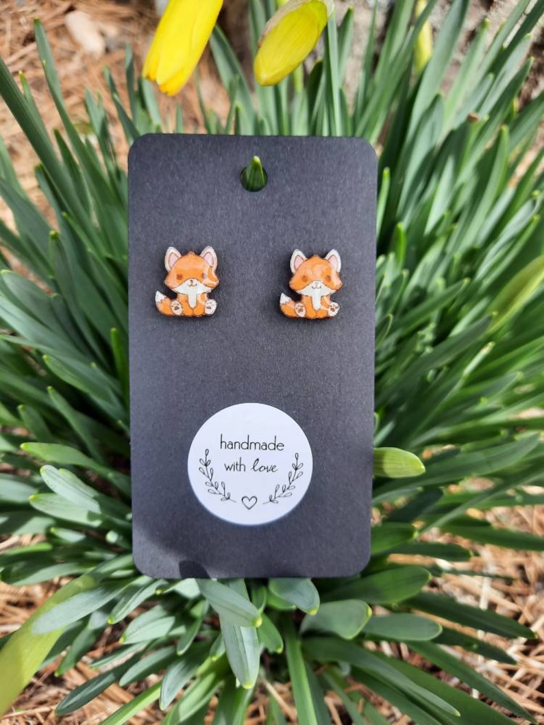 May include: Fox-shaped stud earrings on a black card. The earrings are orange and white with a cartoon design. The card has a small hole at the top and a white sticker that says "handmade with love". The background is green.