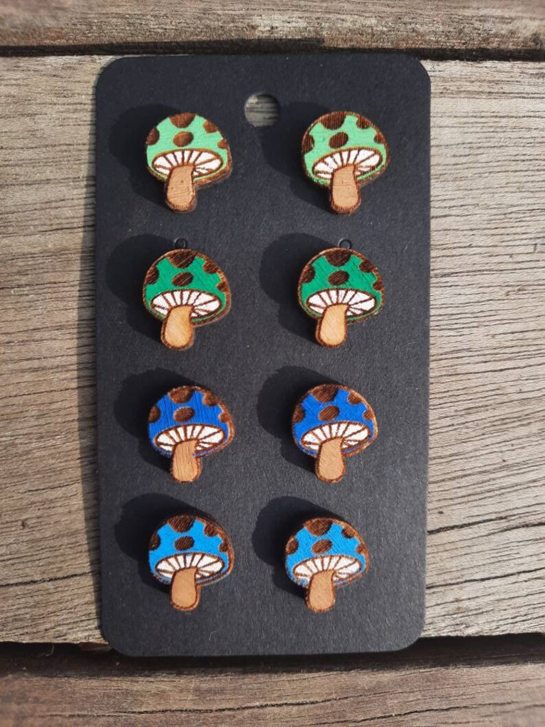 May include: Six pairs of wooden mushroom earrings. Three pairs are green with white polka dots and three pairs are blue with white polka dots.