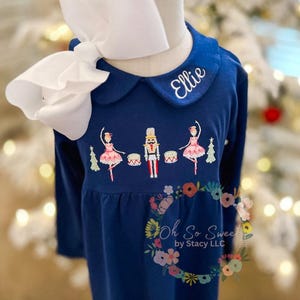 May include: A navy blue long-sleeve shirt with a white Peter Pan collar embroidered with the name Ellie. The chest features an embroidered scene of two ballerinas, a nutcracker, two drums, and two Christmas trees. A large white hair bow is attached.
