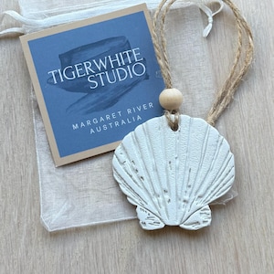 May include: A white seashell ornament with a distressed finish, suspended by a jute string and wooden bead. The ornament is accompanied by a blue tag reading "TIGERWHITE STUDIO MARGARET RIVER AUSTRALIA" and a sheer bag.
