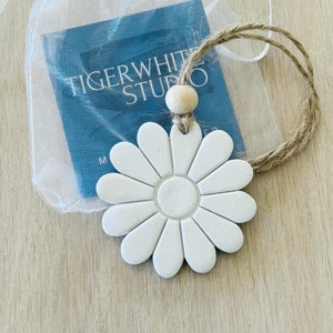 Könnte beinhalten: Ein weißes, gänseblümchenförmiges Ornament mit einer Juteschlaufe und einer Holzperle, präsentiert in einem durchsichtigen weißen Beutel mit einer blauen Karte. Die Karte zeigt den Text "TIGERWHITE STUDIO". Das Ornament befindet sich auf einer hellen Holzoberfläche.