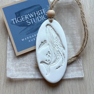 Op de afbeelding: Wit ovaalvormig ornament met een walvisontwerp, een houten kraal en een jute touwtje. Het ornament zit in een transparant zakje met een blauw label met de tekst "TIGERWHITE STUDIO". Het ornament ligt op een licht houten oppervlak.