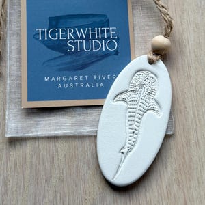 May include: A white oval ceramic ornament featuring an embossed whale shark design, suspended by a jute twine cord with a wooden bead. It is accompanied by a square card labelled TIGERWHITE STUDIO MARGARET RIVER AUSTRALIA and a sheer drawstring pouch.