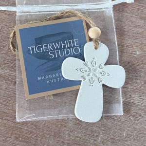 May include: A white ceramic cross ornament with a floral design, suspended by a jute string and wooden bead. The ornament is packaged in a clear drawstring bag with a blue tag reading "TIGERWHITE STUDIO MARGARET AUSTRA".
