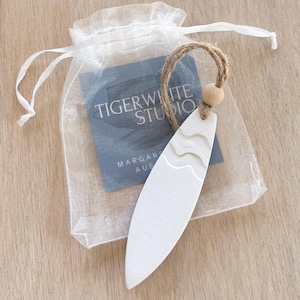 以下が含まれることがあります： 波の模様が施された白いセラミック製のサーフボードのオーナメント。白いオーガンザの袋に、茶色の紐と木製のビーズで包装されています。袋には、「Tigerwhite Studio, Margaret River, Australia.」と書かれたカードも入っています。