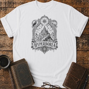 Puede incluir: Una camiseta blanca de manga corta con un gráfico en estilo de tinta negra que muestra un castillo en la cima de una montaña. El diseño incluye una brújula, un cuervo, un oso y el texto "HYPERBOREA" centrado en un marco decorativo con carámbanos.