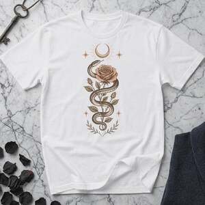 Può includere: T-shirt bianco con un'illustrazione dettagliata di un serpente avvolto attorno a una rosa. Il design include una luna crescente, stelle ed elementi botanici nei toni del marrone e pesca. Una chiave e tessuto scuro sullo sfondo.