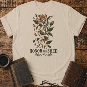 Può includere: Una t-shirt beige a maniche corte con un'illustrazione grafica di un serpente avvolto attorno a una pianta fiorita con foglie. Sotto l'illustrazione, il testo recita HONOR THE SHED in lettere maiuscole di colore verde scuro.
