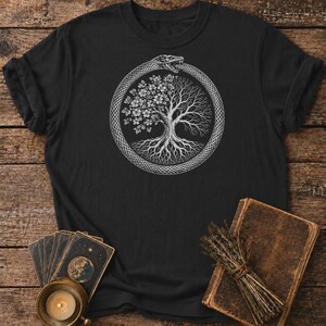 Könnte beinhalten: Ein schwarzes Kurzarm-T-Shirt mit einem weißen Grafikdruck einer Ouroboros-Schlange, die einen Baum umschlingt. Der Baum ist so dargestellt, dass eine Hälfte in voller Blüte steht, während die andere Hälfte kahl mit sichtbaren Ästen ist.