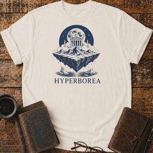 Puede incluir: Una camiseta de color crema con un gráfico azul oscuro de una isla flotante con un templo clásico, montañas y una luna llena. Debajo de la ilustración, la palabra HYPERBOREA está impresa en una fuente serif azul oscuro en mayúsculas.