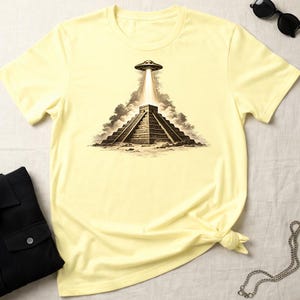 May include: Pale yellow t-shirt featuring a graphic of a pyramid with a UFO above it, emitting a beam of light. The design is in shades of brown and gray, creating a vintage or retro aesthetic. The shirt is a casual wear item.