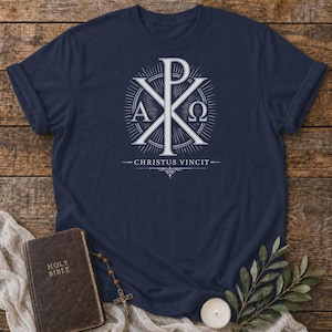 Peut inclure: Un t-shirt bleu marine arborant un motif blanc représentant le symbole du Chi-Rho avec les lettres grecques Alpha et Oméga de chaque côté. Sous le symbole, le texte CHRISTUS VINCIT est écrit en police serif, orné d'une petite fioriture.