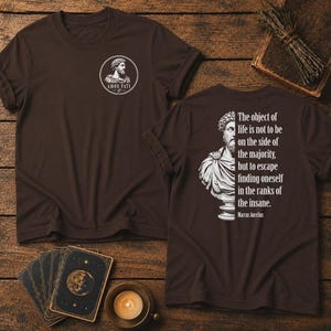 Pode incluir: Uma t-shirt castanho-escura com um gráfico circular branco no peito com um busto de Marco Aurélio e o texto Amor Fati. As costas apresentam um gráfico branco de um busto e a citação The object of life is not to be on the side of the majority, but to escape finding oneself in the ranks of the insane.