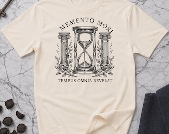 May include: Cream-colored t-shirt featuring a black hourglass design between two pillars, with the words "MEMENTO MORI" and "TEMPUS OMNIA REVELAT" printed above and below the hourglass. The design has a vintage, classical aesthetic.
