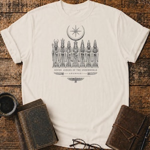 Puede incluir: Una camiseta de color crema con un gráfico en tinta negra de siete figuras con túnicas y tocados ornamentados. Arriba hay una luna creciente y una estrella. Debajo de las figuras, el texto dice: SEVEN JUDGES OF THE UNDERWORLD ANUNNAKI.