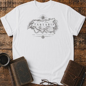 Op de afbeelding: Een wit T-shirt met korte mouwen en een zwarte inktstijl grafische afbeelding van een kaart van Eurazië. De tekst TARTARIA staat boven de kaart, met THE FORGOTTEN EMPIRE eronder, vergezeld van een kompasroos en een zon-icoonontwerp.