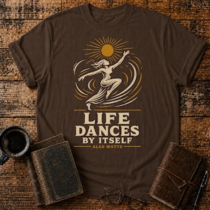 Może przedstawiać: Brązowa koszulka z graficznym wzorem przedstawiającym tańczącą postać, słońce i napis "LIFE DANCES BY ITSELF - ALAN WATTS" w kolorze białym i złotym. Koszulka jest prezentowana na drewnianej powierzchni z książkami i filiżanką kawy.