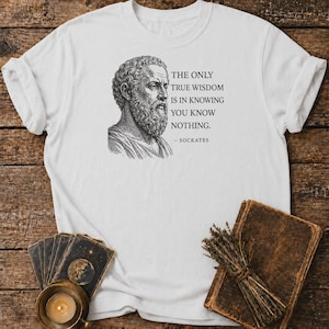 Könnte beinhalten: Ein weißes T-Shirt mit einer schwarzen Illustration im Tintenstil des Philosophen Sokrates. Rechts neben dem Porträt steht der Text: THE ONLY TRUE WISDOM IS IN KNOWING YOU KNOW NOTHING. - SOCRATES. Das T-Shirt hat kurze Ärmel und einen Rundhalsausschnitt.