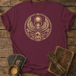 May include: Burgundy t-shirt featuring a detailed gold illustration of an Egyptian scarab beetle with wings, a sun, and other hieroglyphic elements. The design is centered on the shirt. The shirt is displayed on a wooden surface with tarot cards, a candle, and a book.