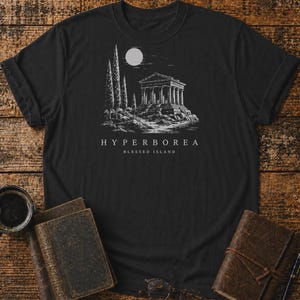 Puede incluir: Una camiseta negra de manga corta con un estampado gráfico en blanco que muestra un templo clásico, cipreses altos y una luna llena. Debajo de la ilustración, el texto dice HYPERBOREA BLESSED ISLAND en una fuente sans-serif.